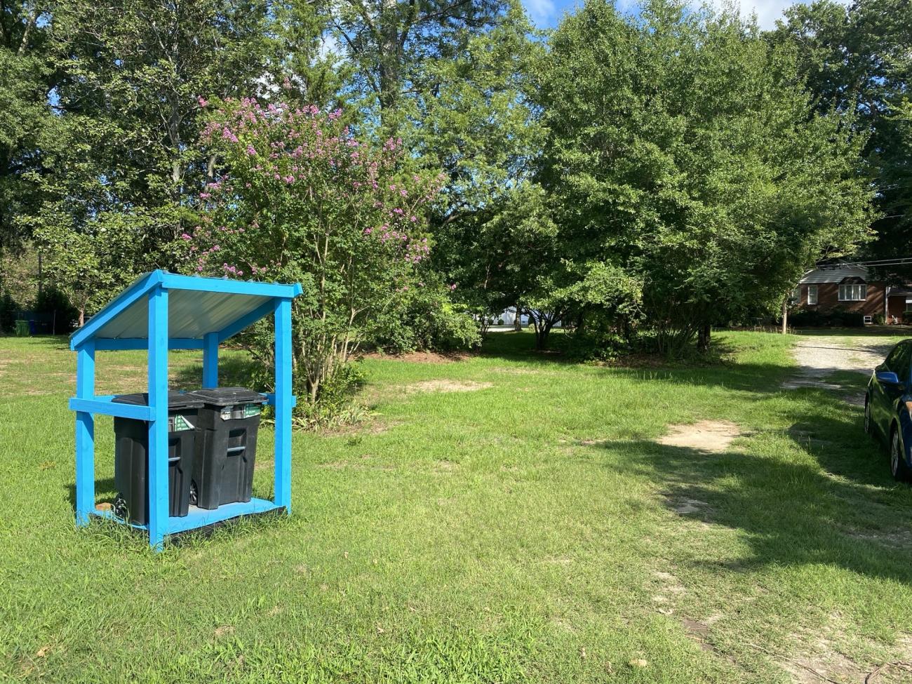 Bright blue shelter holding two trash carts beside a grassy driveway surrounded by trees.