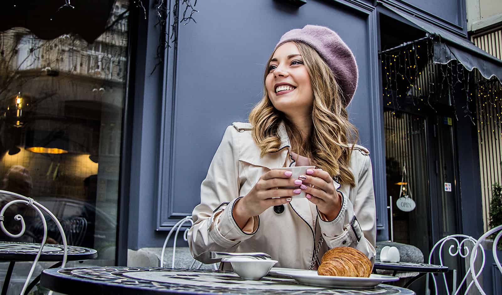 Cheerful french woman. Low-angle view of beautiful blonde woman in beret looking away and smiling while sitting in french vintage cafe. Woman drinking coffee with croissant. French breakfast concept.