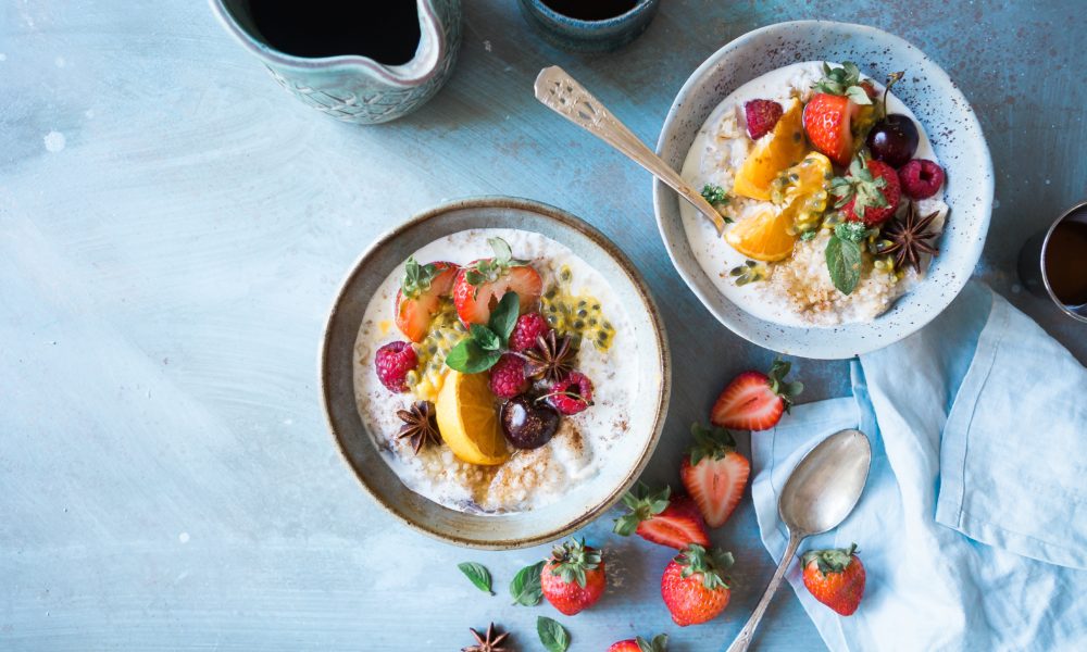 two small bowls of oatmeal topped with oranges, grapes, berries, seeds, and green leaves beside are 7 slices of strawberries, spoon, and blue cloth blue background, starchy foods for a slim body, Starchy Foods You can Eat to Stay Slim