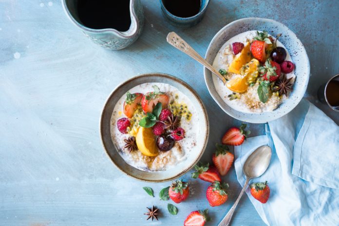 two small bowls of oatmeal topped with oranges, grapes, berries, seeds, and green leaves beside are 7 slices of strawberries, spoon, and blue cloth blue background, starchy foods for a slim body, Starchy Foods You can Eat to Stay Slim