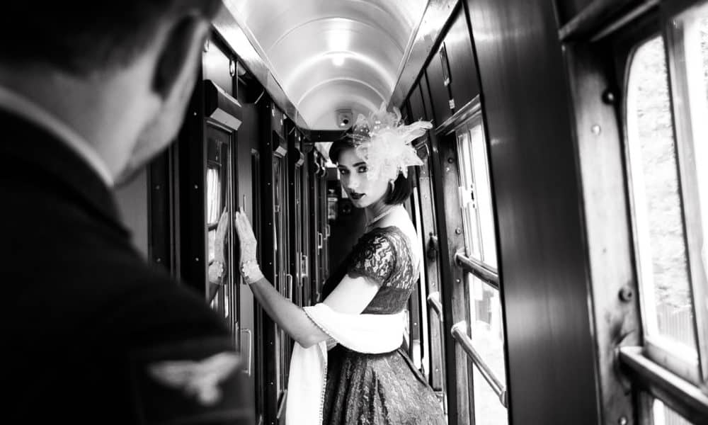 Portrait of beautiful woman in vintage tea dress standing in corridor of locomotive train as air force officer in uniform watches her