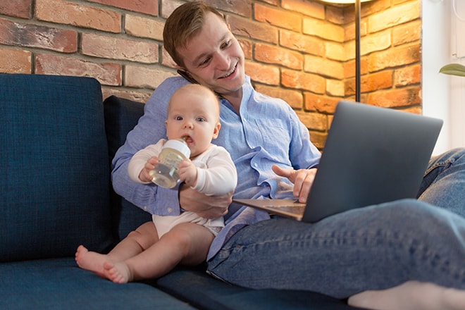 Dad works on a laptop at home holding a baby in his arms feeding it from a bottle with a pacifier. Dad on maternity leave earns money online